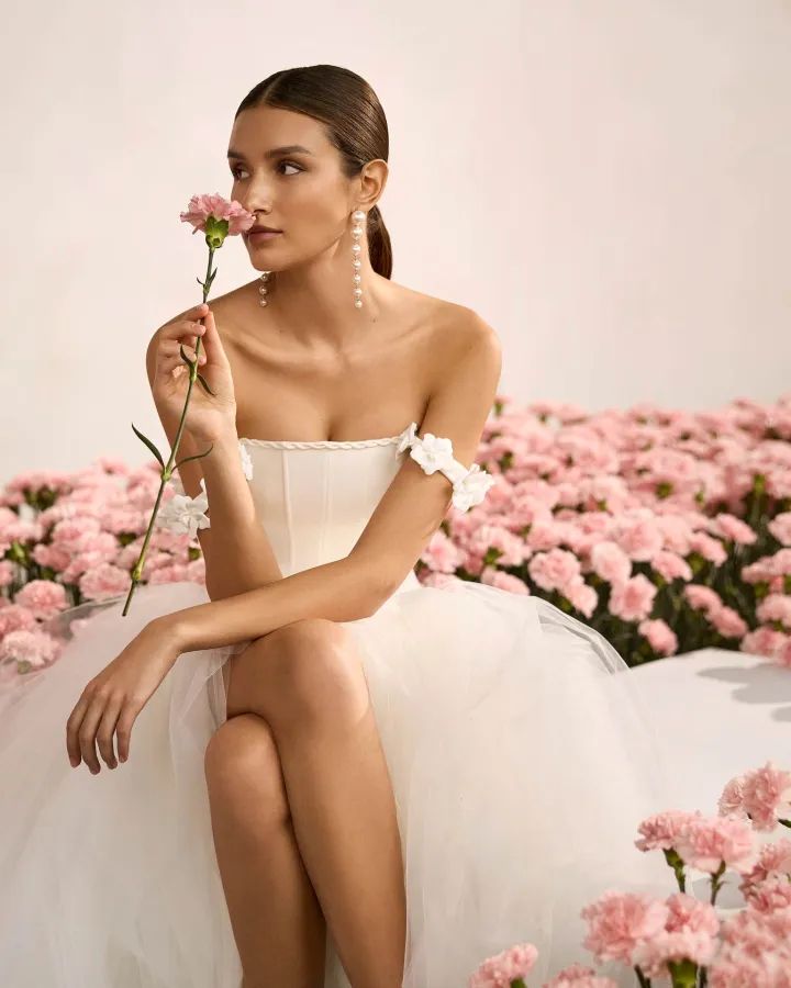 Model mit Rosen sitzt auf dem Bett mit einem Kleid aus der Rosa Clará Kollektion 2026