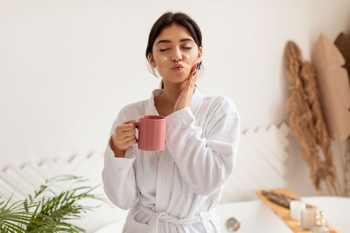 JungJunge Frau mit einer Tasse Tee cremt sich das Gesicht im Zimmere Frau cremt sich das Gesicht im Zimmer mit einer Tasse Tee