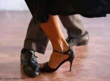 Close-up of a woman in a black strappy high heel stepping near a man's black dress shoe on a wooden dance floor.