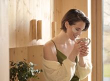 Woman in a green dress and cream cardigan relaxing in a wooden room, smiling while sipping from a mug; she has eye patches under her eyes and sits near leafy plants by a window.