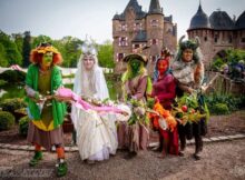 Group of six people in colorful fantasy costumes with flower garlands, posing in front of Burg Satzvey castle during a festival.