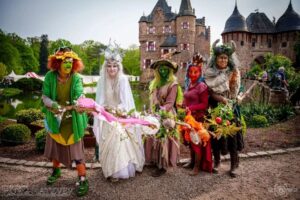 Group of six people in colorful fantasy costumes with flower garlands, posing in front of Burg Satzvey castle during a festival.