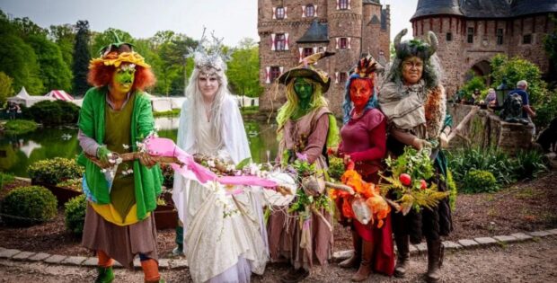 Group of six people in colorful fantasy costumes with flower garlands, posing in front of Burg Satzvey castle during a festival.