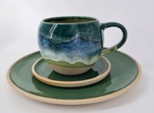 Green ceramic mug with blue glaze sits on a matching green plate and beige saucer stack.