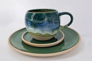 Green ceramic mug with blue glaze sits on a matching green plate and beige saucer stack.