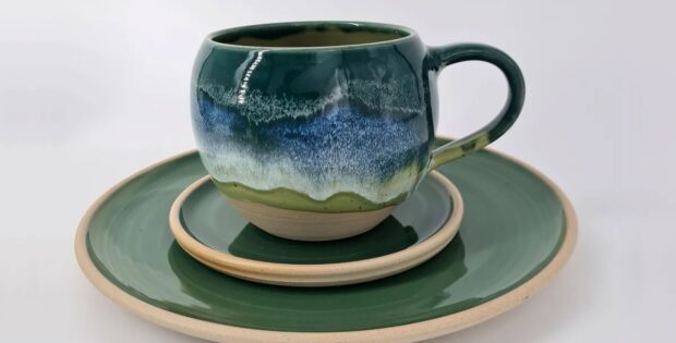 Green ceramic mug with blue glaze sits on a matching green plate and beige saucer stack.