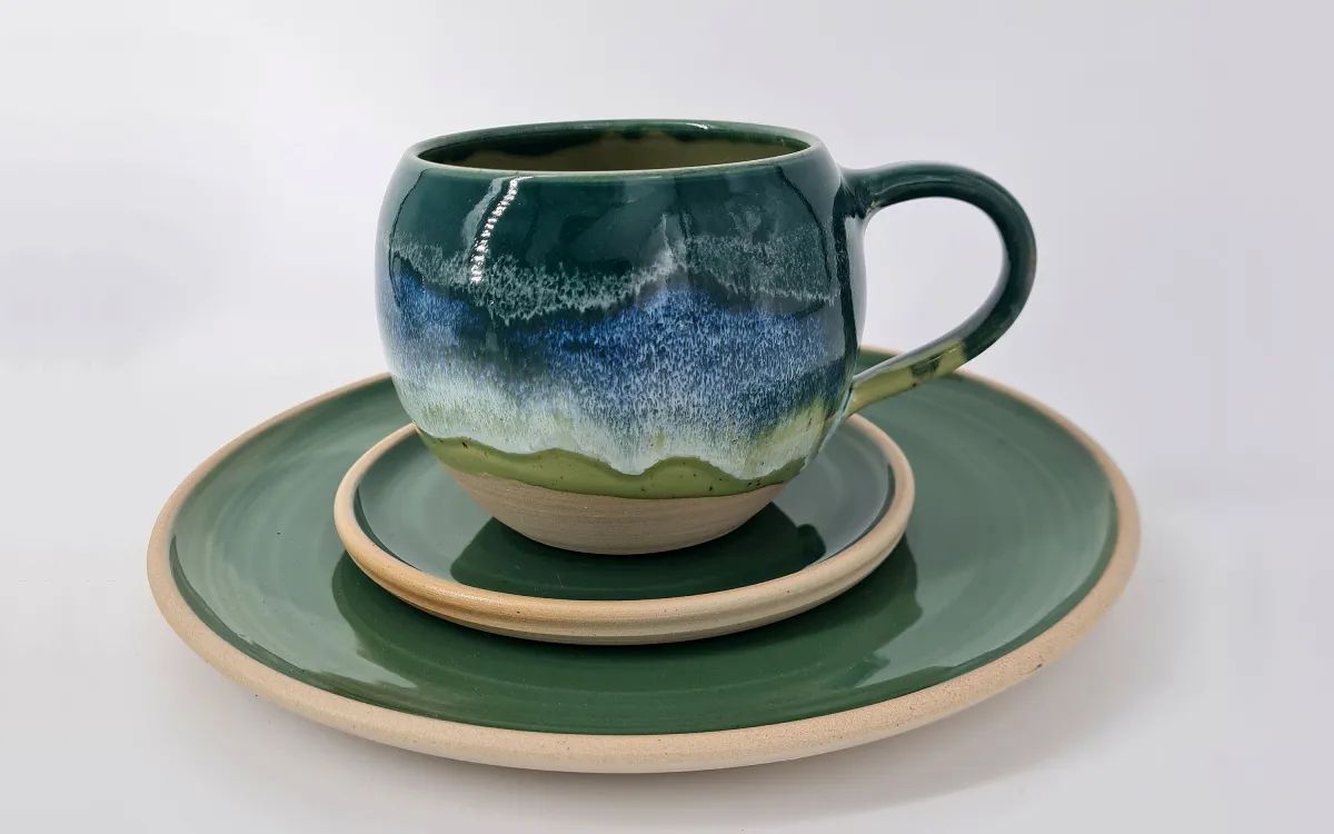 Green ceramic mug with blue glaze sits on a matching green plate and beige saucer stack.