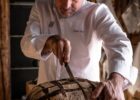 Bald chef in a white coat slices a round crusty loaf with a knife in a rustic bakery setting of wood walls and tools behind him.