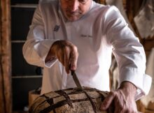 Bald chef in a white coat slices a round crusty loaf with a knife in a rustic bakery setting of wood walls and tools behind him.