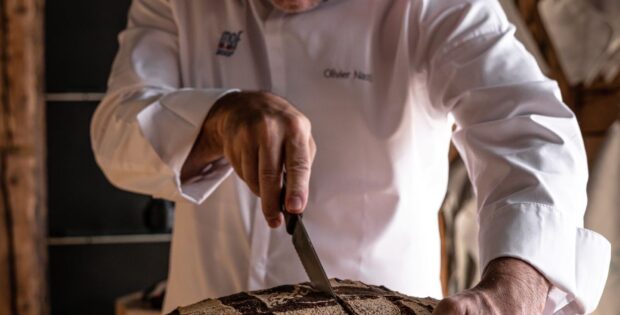 Bald chef in a white coat slices a round crusty loaf with a knife in a rustic bakery setting of wood walls and tools behind him.