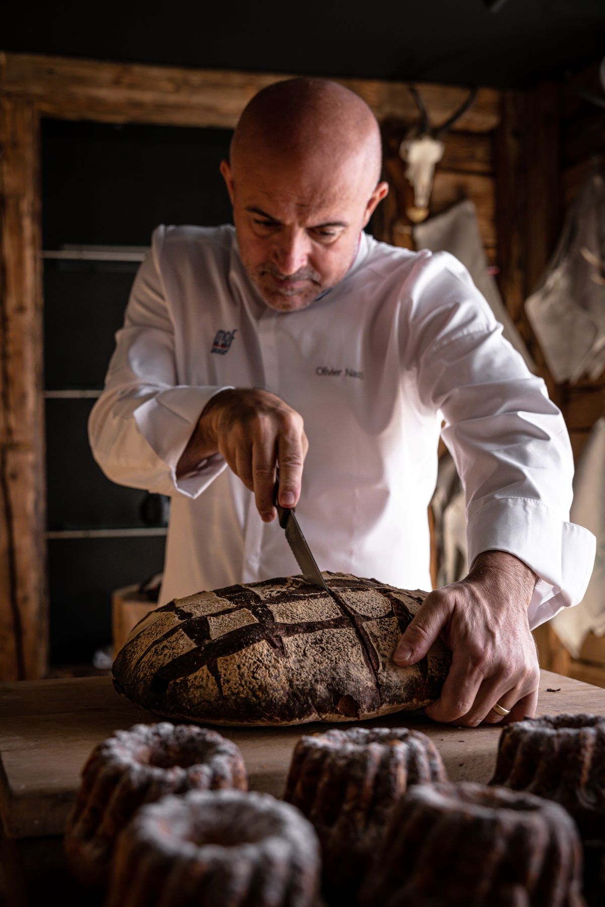 Bald chef in a white coat slices a round crusty loaf with a knife in a rustic bakery setting of wood walls and tools behind him.